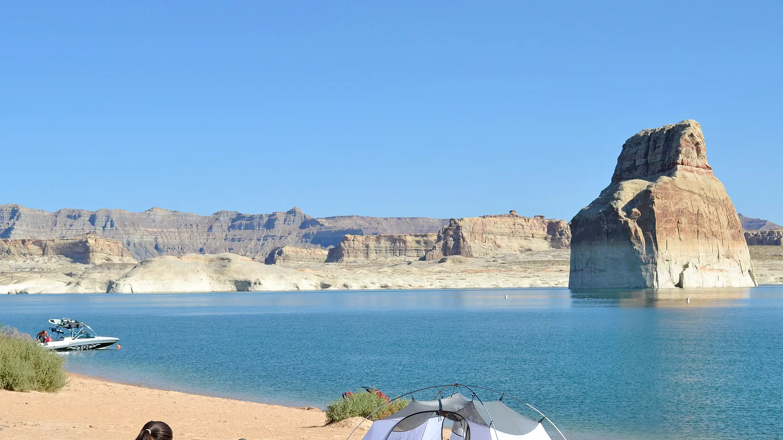 Lone Rock standing in Lake Powell off Lone Rock Beach, with a tent and a boat near the shore