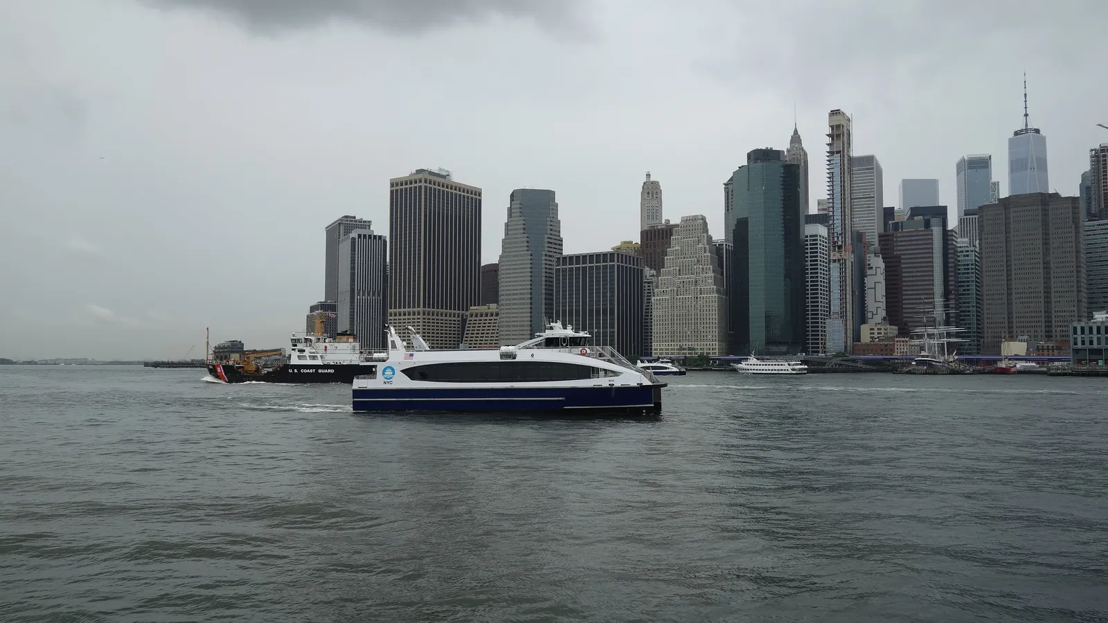 An NYC Ferry crossing the East River in front of Lower Manhattan, seen from Brooklyn Bridge Park