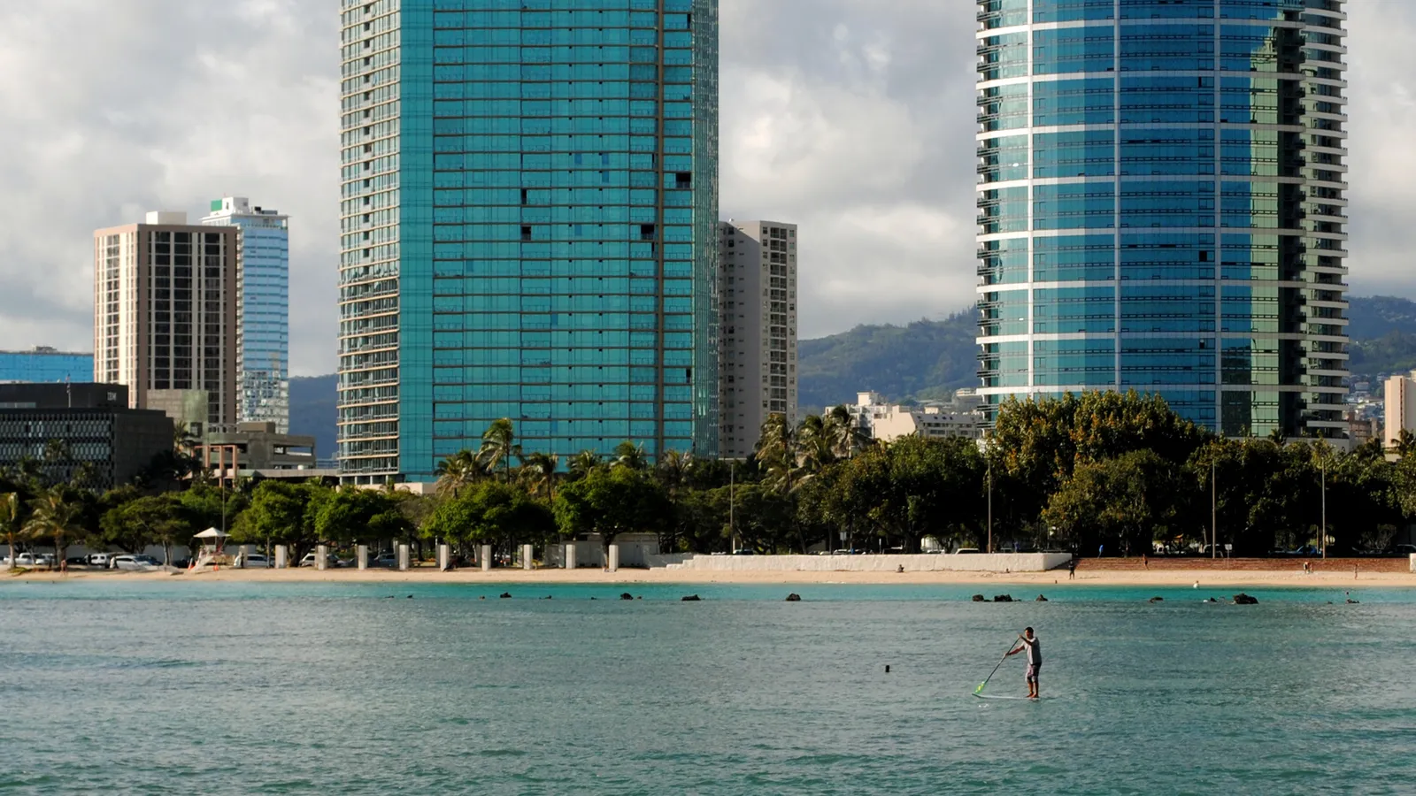 A stand-up paddler on calm water off Ala Moana Beach Park, with Honolulu high-rises behind