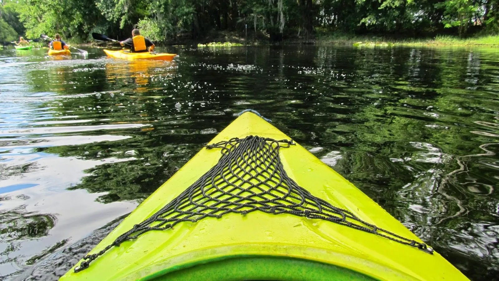 Kayakers paddling a shaded stretch of Shingle Creek, seen from the bow of a kayak