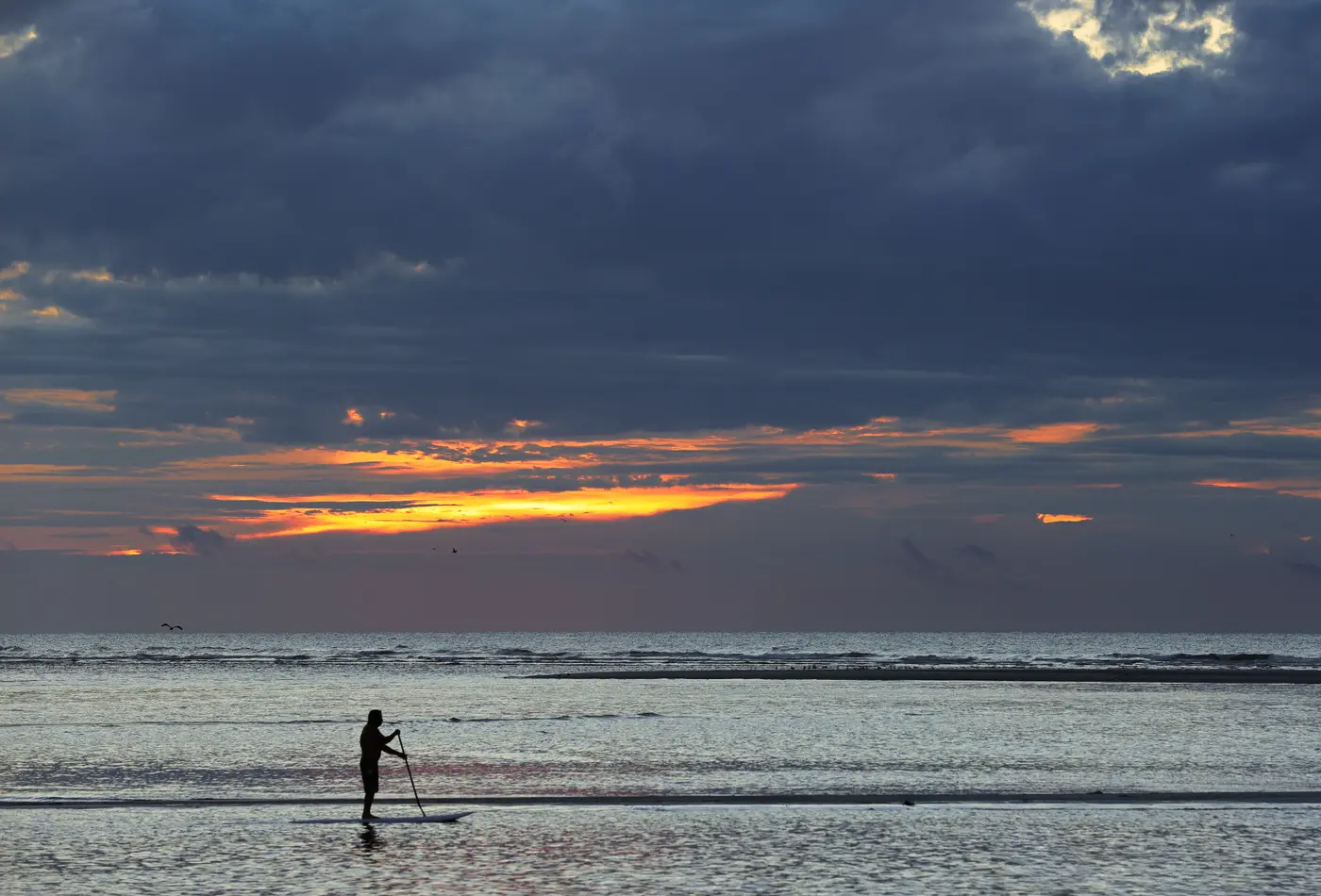 Paddler standing on a board in shallow water under dark clouds at sunrise
